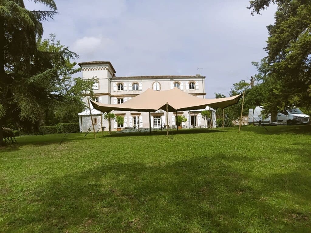 Grande tente stretch beige sur une pelouse verte devant un élégant château. Arbres luxuriants et vans indiquent des préparatifs événementiels en extérieur.
