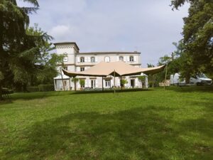 Grande tente stretch beige sur une pelouse verte devant un élégant château. Arbres luxuriants et vans indiquent des préparatifs événementiels en extérieur.