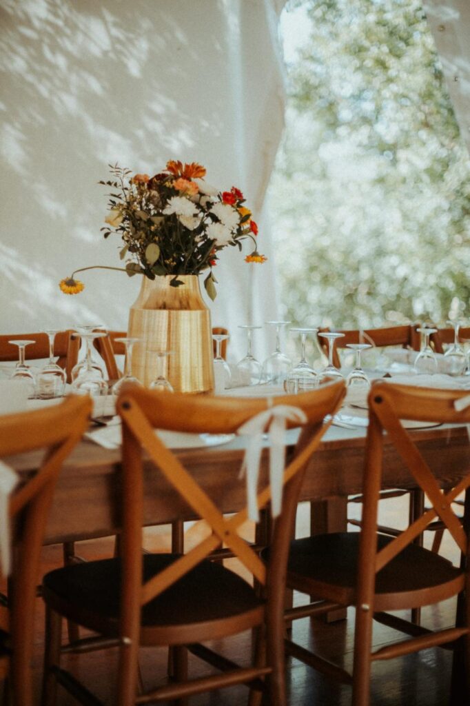 Table de réception élégante avec vase doré de fleurs vives, verres alignés et chaises en bois, sous une tente ombragée.