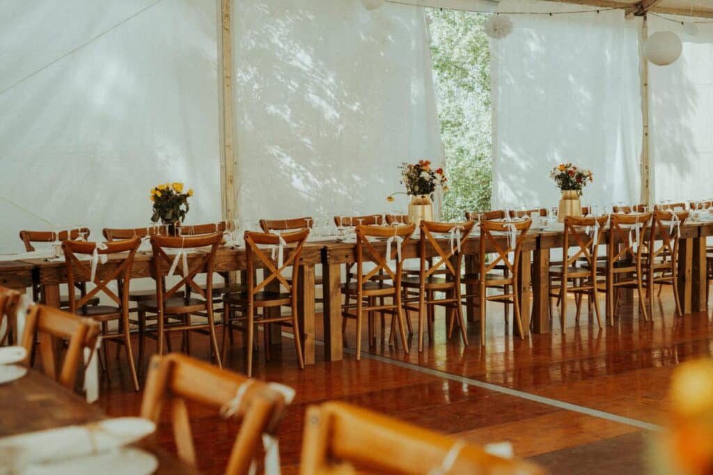 Longue table en bois et chaises rustiques avec rubans blancs pour une réception sous tente. Vases de fleurs colorées, vaisselle prête, sol en bois brillant. Ombres de feuilles sur la toile.